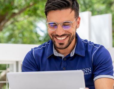 male student wearing a Lynn University polo shirt smiling and working on a laptop outdoors